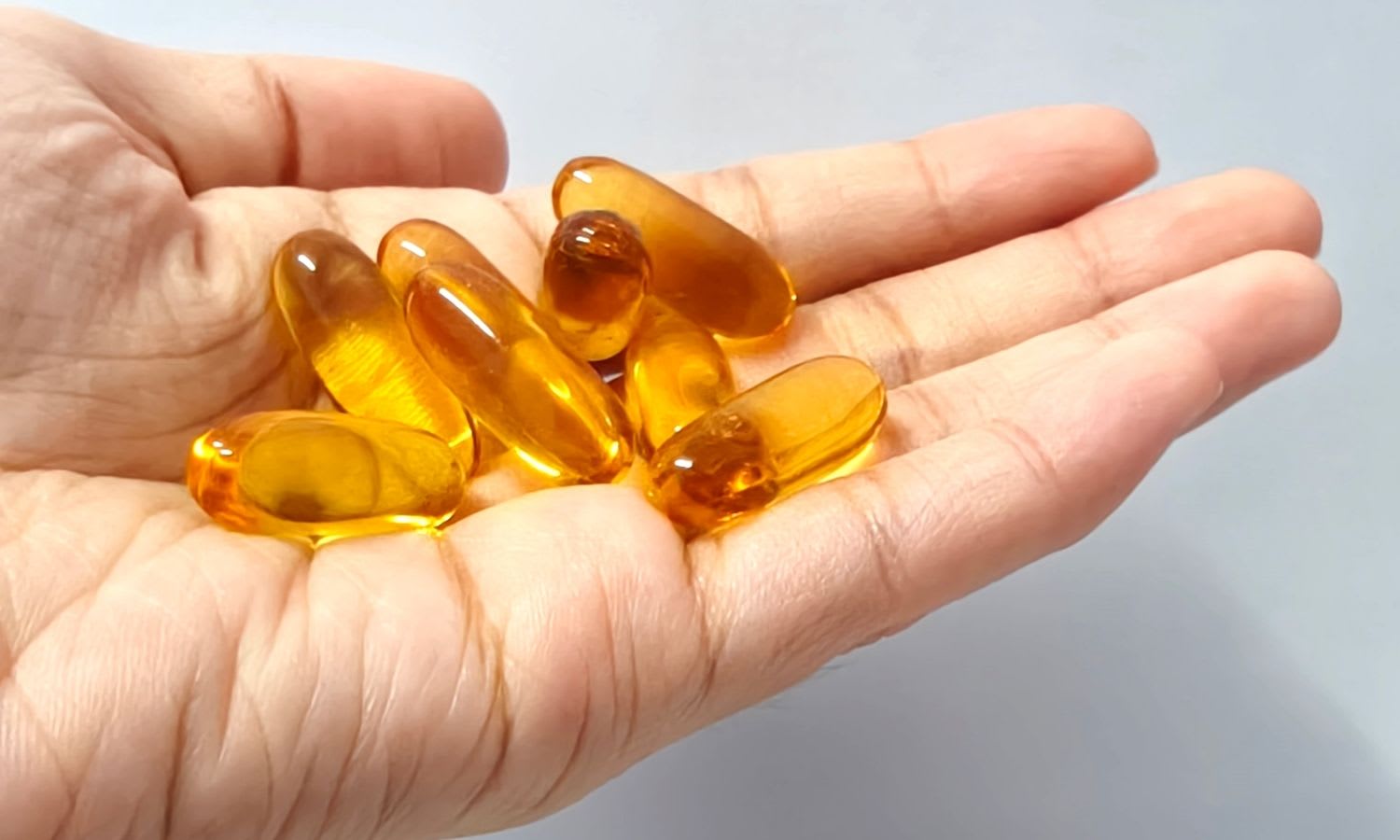 A hand holds several yellow, translucent softgel capsules—an example of omega-3 supplement quality—against a plain light background.